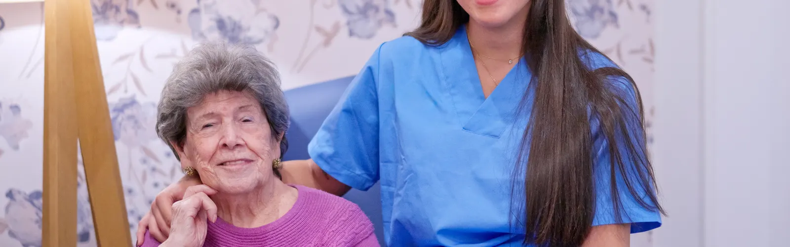 Cuidadora sentada junto a una mujer mayor, las dos de frente y sonriendo