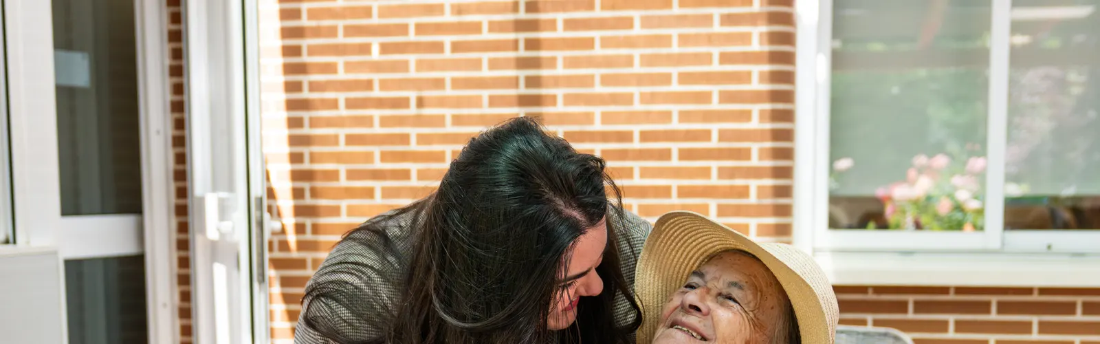 Cuidadora abrazando a una mujer mayor con sombrero de paja en una terraza